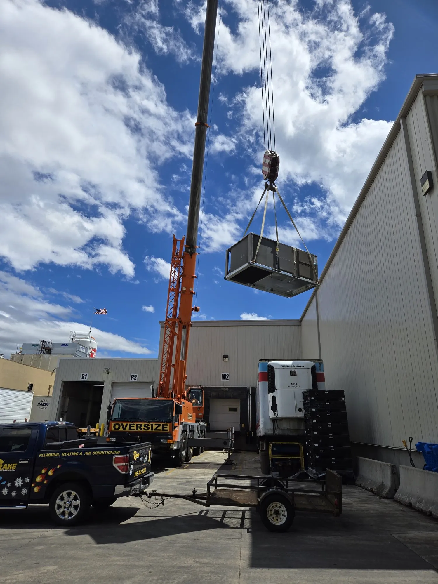 A crane lifting a rooftop HVAC unit onto a commercial building, with a Drake Mechanical truck below